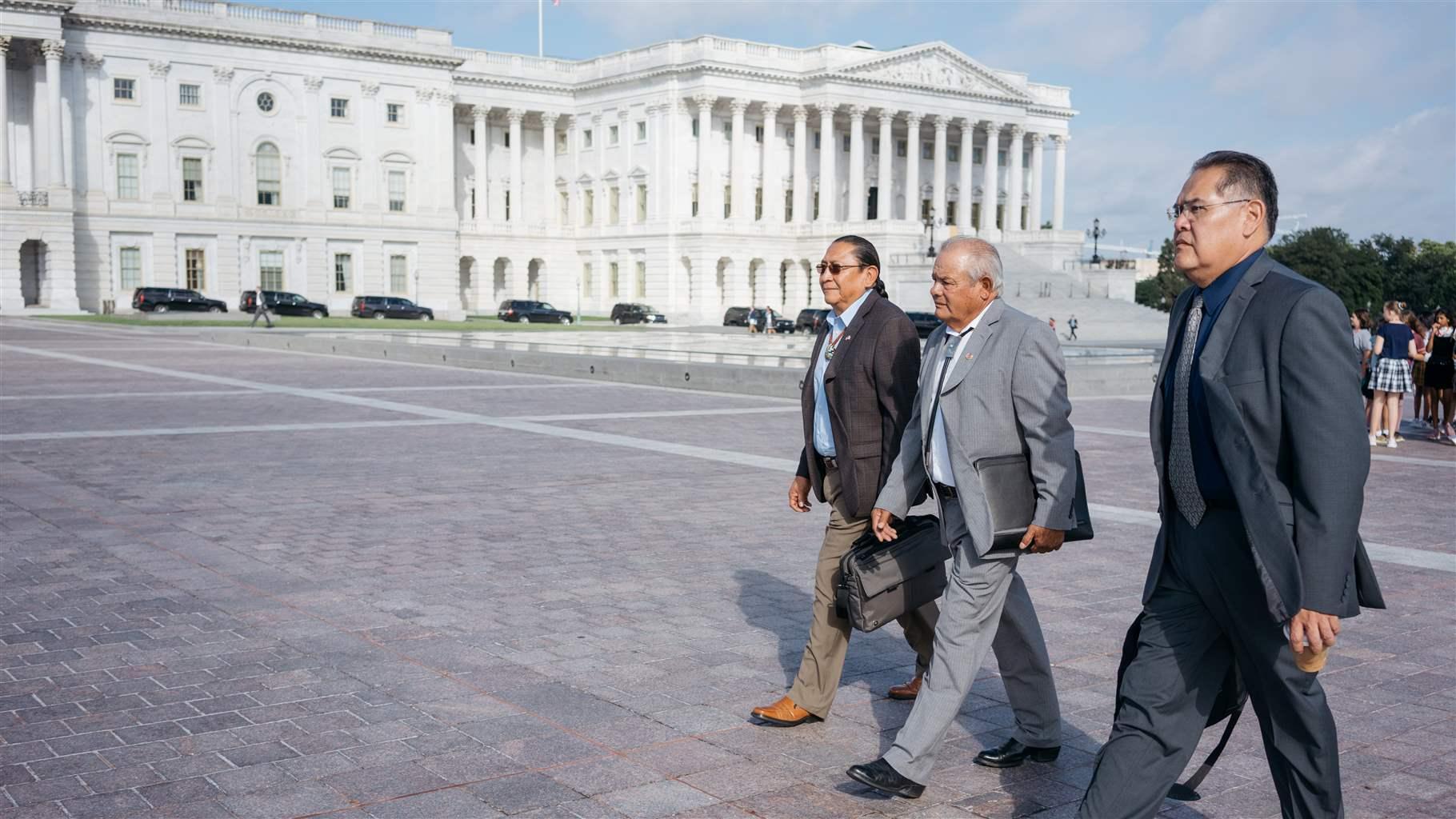 Three men in suits walk across smooth, symmetrical, reddish-gray pavers with the U.S. Capitol, a three-story white marble building, in the background. A line of black SUVs sits in front of the building, the façade of which includes ornate features, including tall columns. 