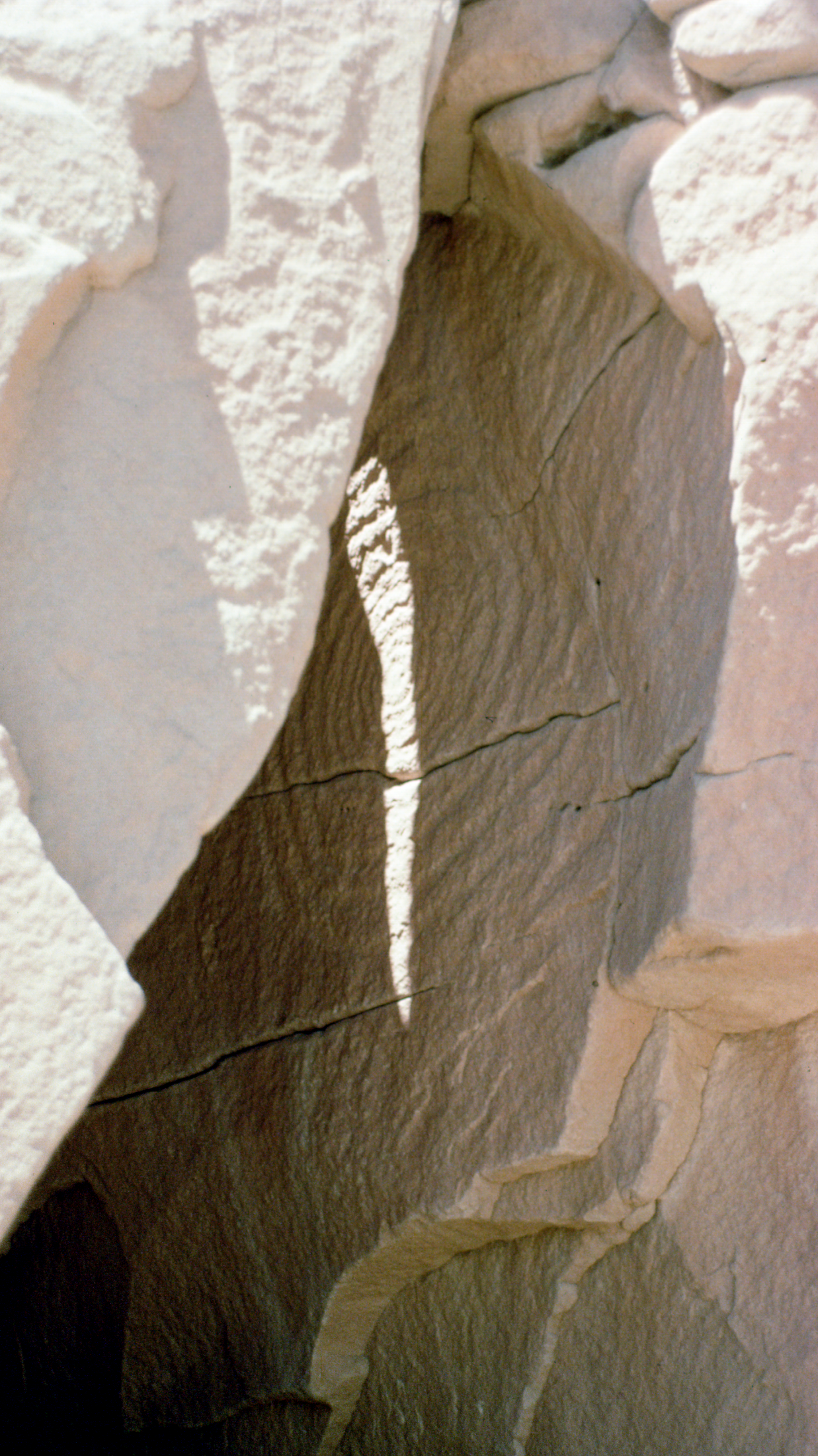 A rock formation with a large, cleaved, cave-like opening that is mostly in shadow except for a vertical sliver of sunlight lighting up the back wall. 