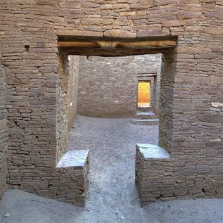 The interior of an ancient stone structure rises from a dirt floor, with one opening in a wall leading to a room and another opening beyond. Natural light appears to come in from above, though no sky or ceiling are visible.