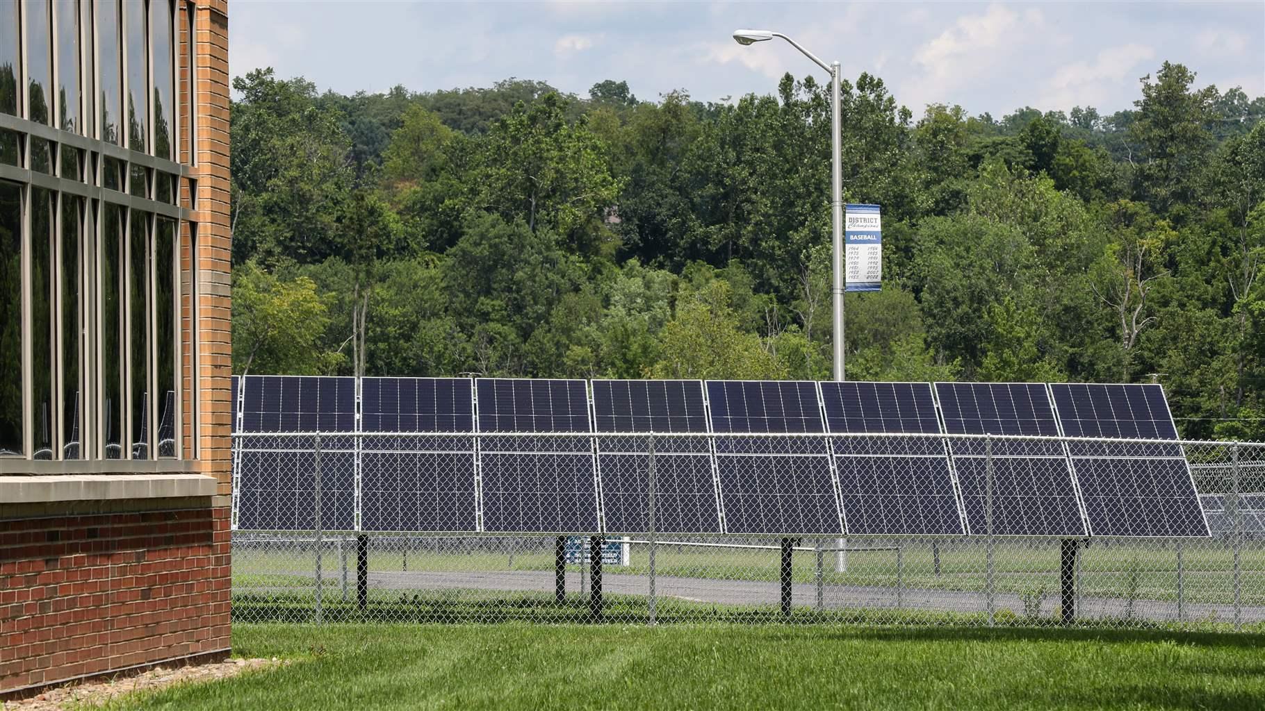 Tilted solar panels stand side by side behind a chain-link fence next to a school building and green field. Beyond the panels, trees rise twice as high as the black-and-silver solar array.