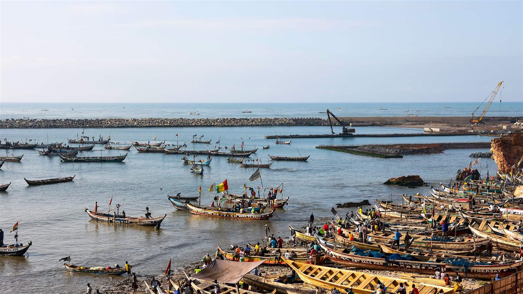 A busy harbor scene, with workers preparing fishing boats on a beach and a dozen or so vessels in the water, including two just off shore on which fishers excitedly wave the Ghanaian flag and another flag.