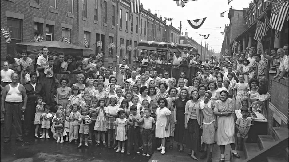 In a black-and-white photo, a crowd of people stands in and along a street with row houses on either side and U.S. flags and bunting hanging overhead.