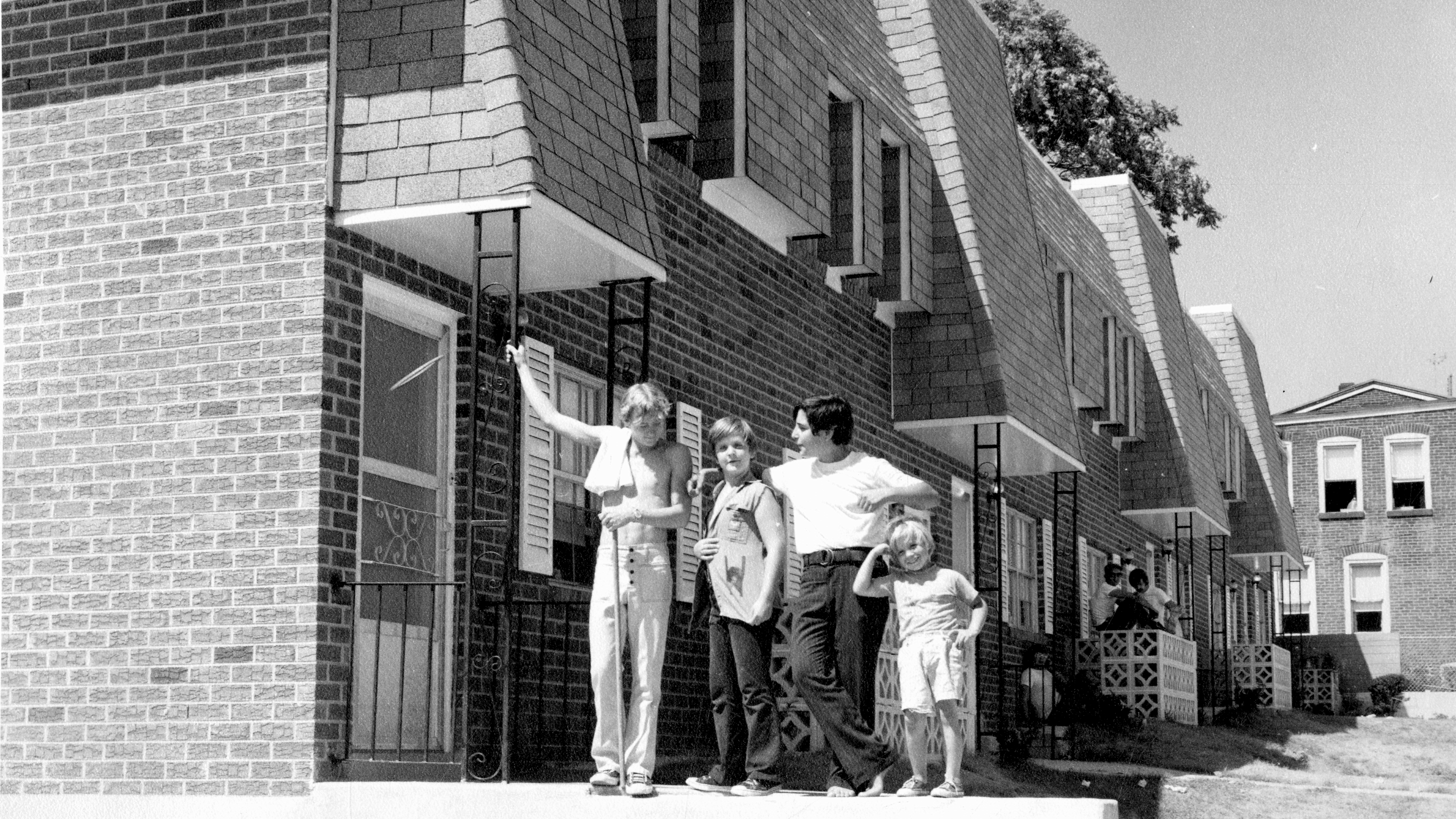 In a black-and-white photo, four people stand together on a sidewalk next to a two-story brick row house with a screen door, shutters, and wrought-iron railings.