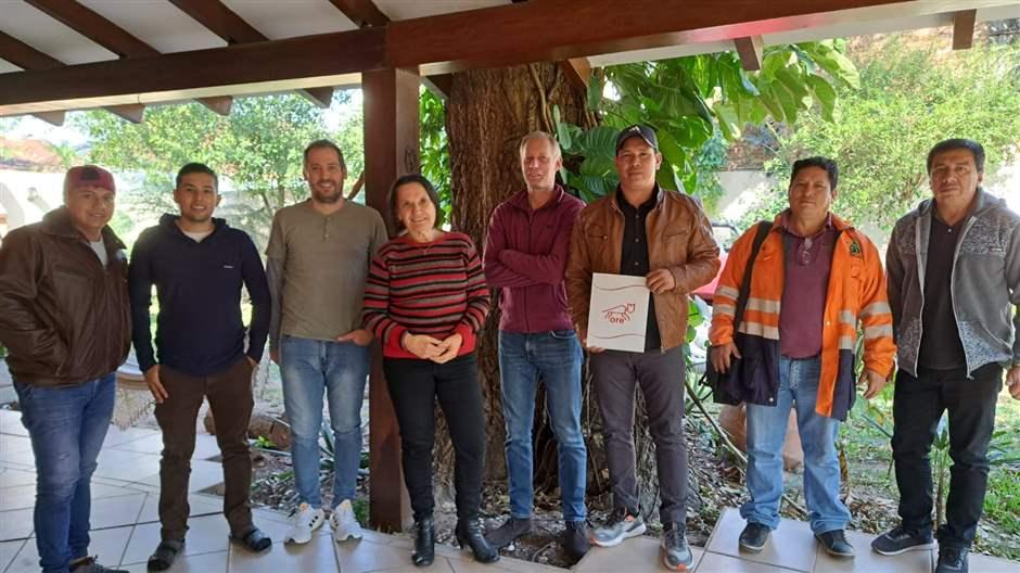 Un grupo de ocho personas—entre ellos Leonardo Tamburini y miembros del gobierno de Charagua—paradas frente a un árbol alto bajo la cubierta de un patio