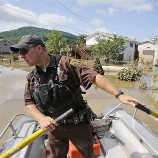 A man in a baseball cap and brown short-sleeved uniform rows a silver boat through muddy floodwaters along a residential street. 