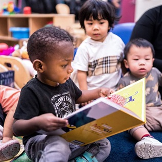 A child reads a hardcover book at Maternity Care Coalition’s Early Head Start child care and early education center in South Philadelphia. In the background, a few other children and an adult sit nearby.