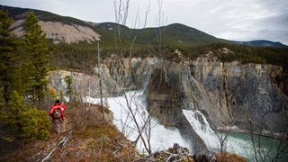 A hiker facing away from the camera and wearing an orange jacket, dark hat, and light brown pants walks on a path toward a rocky overlook near the top of a rushing waterfall. Evergreen trees are at the person's left, and the bare trunks of deciduous trees are at the right.