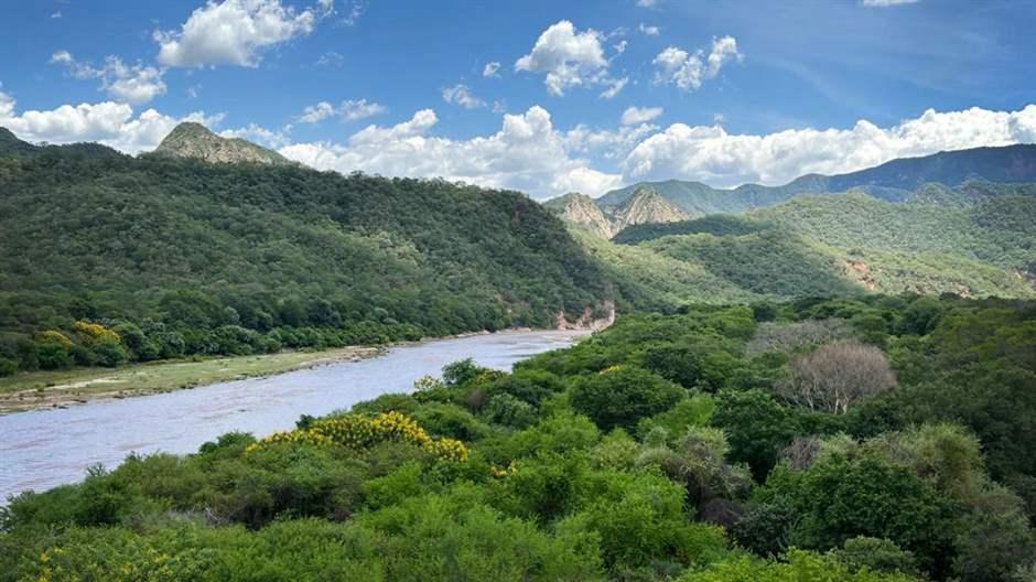 El río Parapetí fluye a través de las montañas y los valles del bosque del Gran Chaco boliviano. Por encima del río, las nubes blancas salpican el cielo azul.   