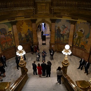 A man in a suit is flanked by colleagues as they pose for a photo at the bottom of a grand staircase. There are colorful paintings of people on the walls in the hall, and other people clad in business outfits watching the photoshoot.