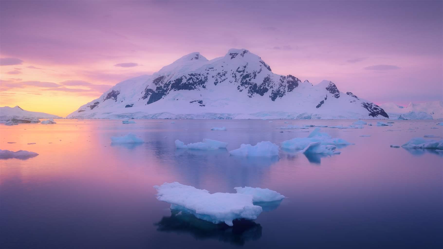 The sun is setting behind a snow-covered mountain, casting an orange glow across still deep blue water. Several ice floes float in the water, and the sky above is a light purple.