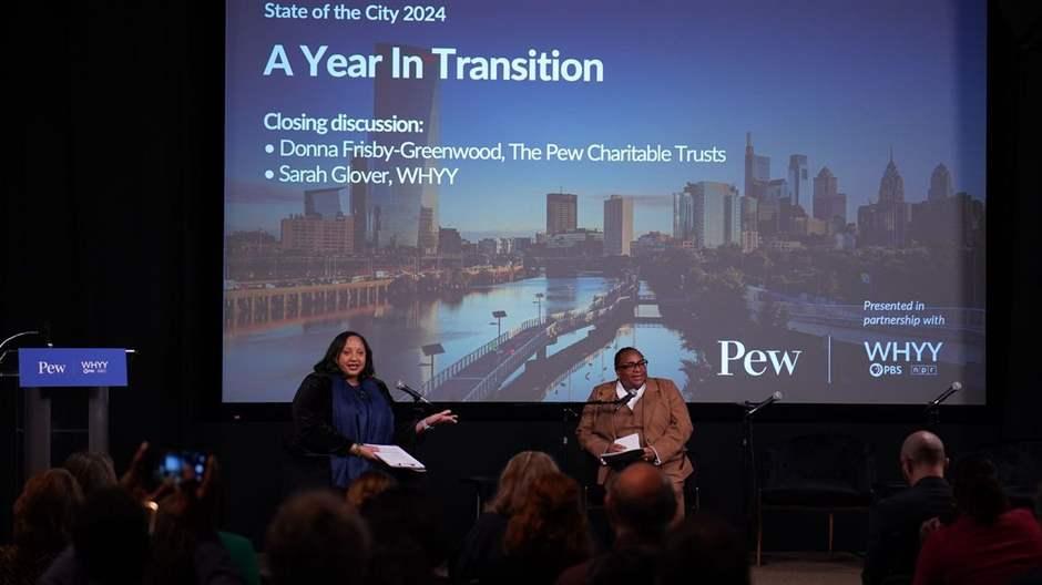 Two women on a stage with microphones have a discussion in front of a live audience. In the background is a large screen with the words “State of the City 2024: A Year in Transition” superimposed over a photo of Philadelphia’s skyline.