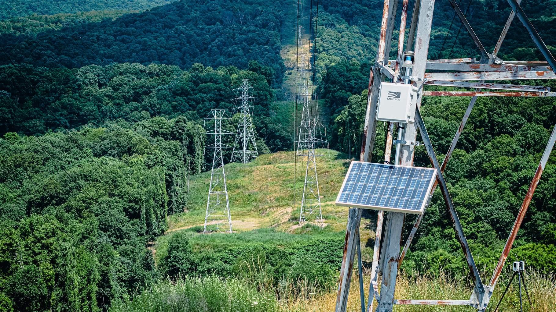 A hilly area covered in green grass and trees with electric transmission towers in the background. In the foreground, a close-up of a panel-like sensor attached to a transmission tower. 