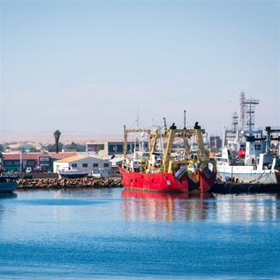 Fishing boats are docked at a port. On the right are two large vessels, the further right vessel is all white with the second red with yellow pillars. Smaller blue vessels are docked on the left side and there is a backdrop of warehouse-style buildings.