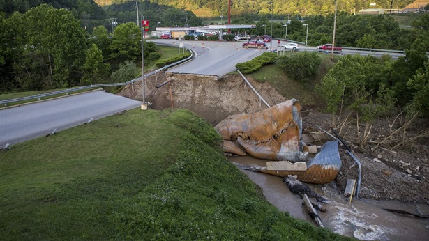 Chunks of a washed-away bridge lie in a riverbed alongside a vast, impassable gap left in a major roadway. A one-story shopping mall sits in the distance, nestled between green hills. 