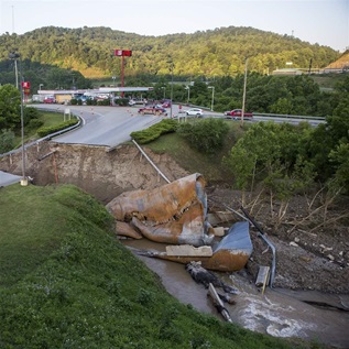 Chunks of a washed-away bridge lie in a riverbed alongside a vast, impassable gap left in a major roadway. A one-story shopping mall sits in the distance, nestled between green hills. 