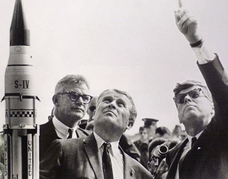 President Kennedy shown pointing at the sky with two other men
