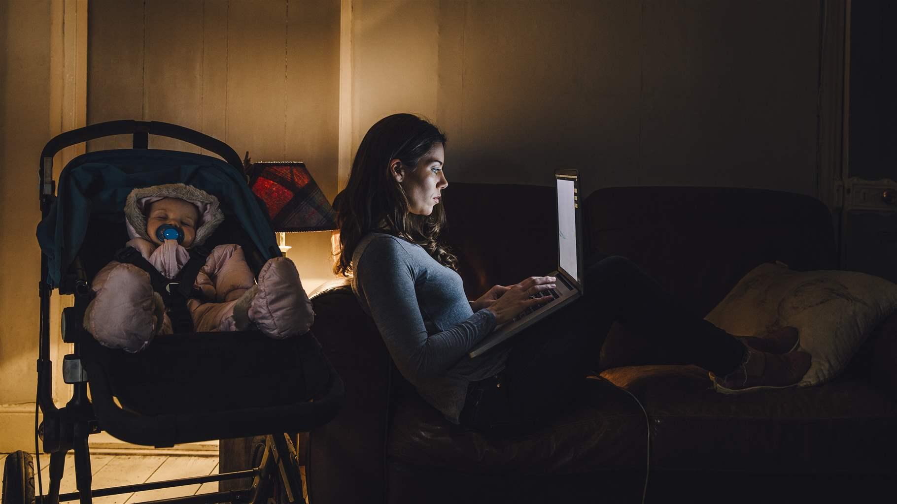 Young mother is working on a laptop on the sofa in her home with her baby daughter sleeping in the pushchair next to her.