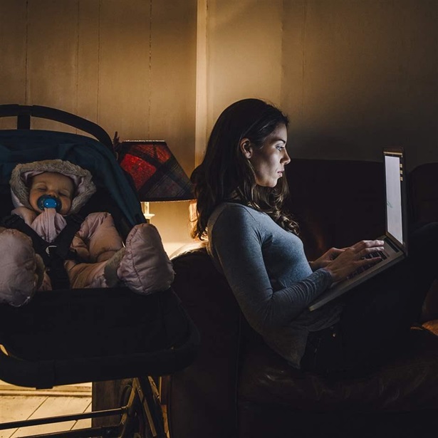 Young mother is working on a laptop on the sofa in her home with her baby daughter sleeping in the pushchair next to her.