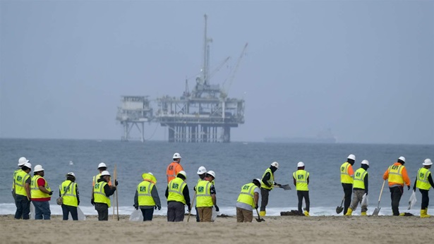 Workers in protective suits clean a contaminated beach with an offshore drilling station in the distance. 