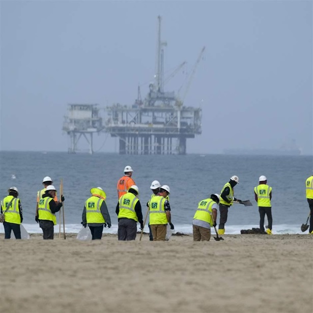 Workers in protective suits clean a contaminated beach with an offshore drilling station in the distance. 