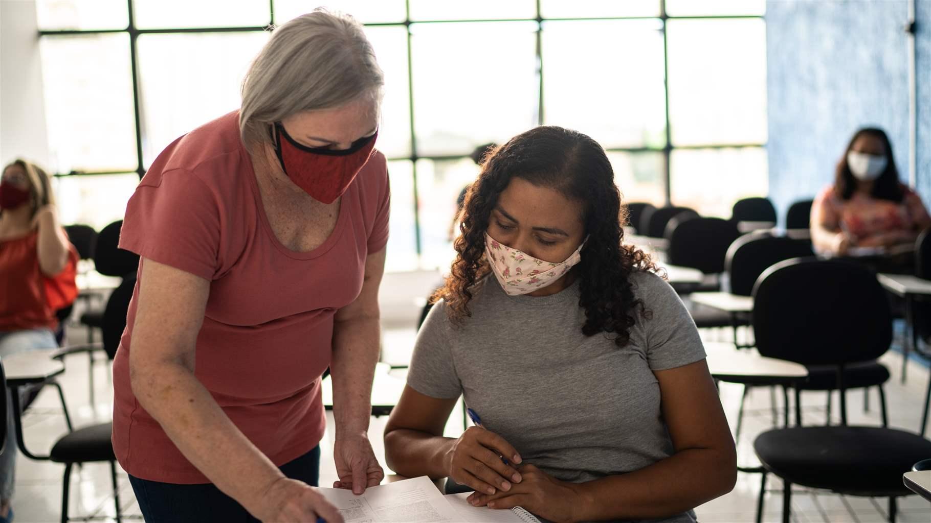 A teach waring a mask looks over a students shoulder to review their paper.