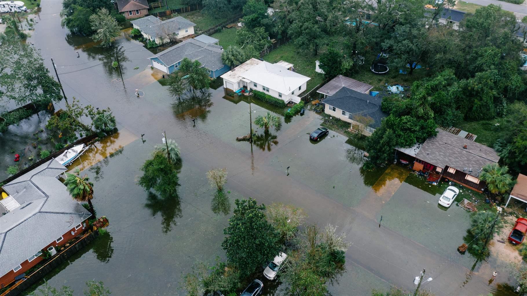 An aerial shot of of a suburban neighborhood with flooded streets and front yards with downed trees and powerlines. 