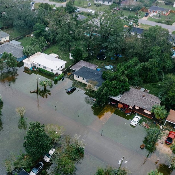An aerial shot of of a suburban neighborhood with flooded streets and front yards with downed trees and powerlines. 