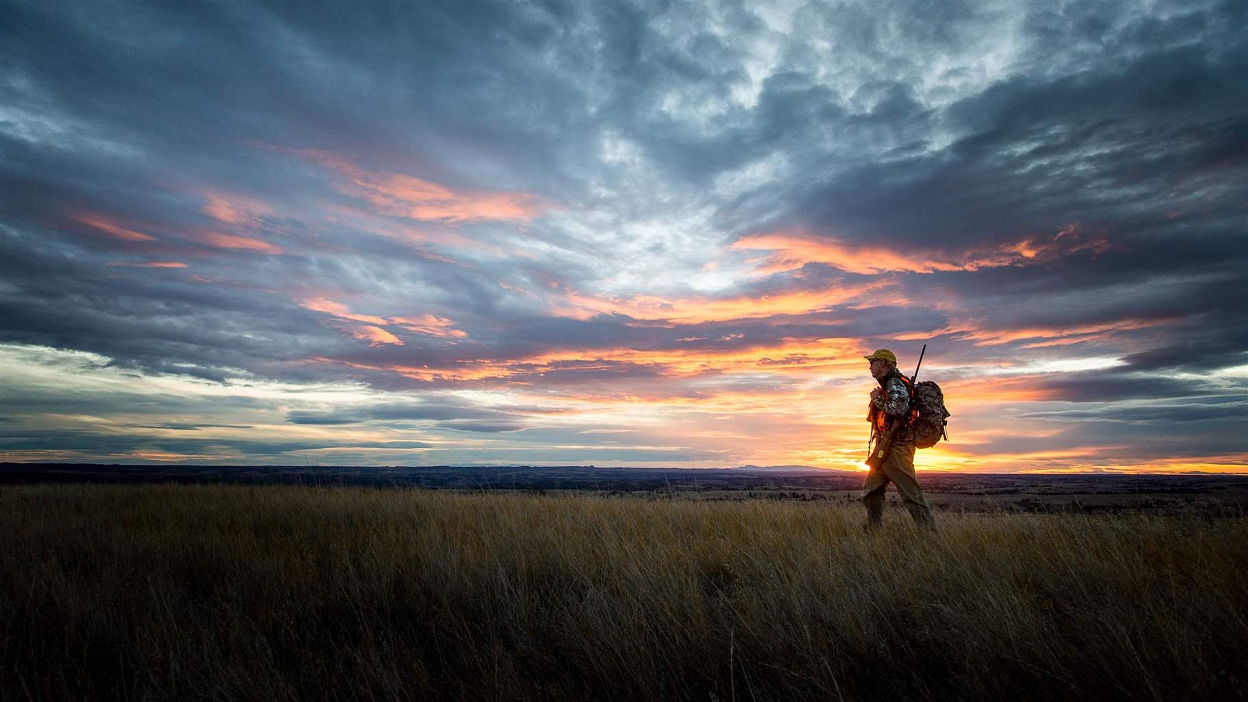 A hunter walking in a field with the sunsetting behind him.