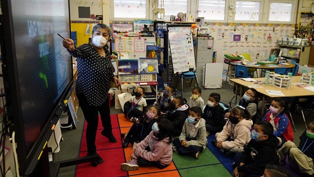 A masked teach points to the a digital screen with a full class of kindergarteners sitting on the ground in front of her wearing masks. 