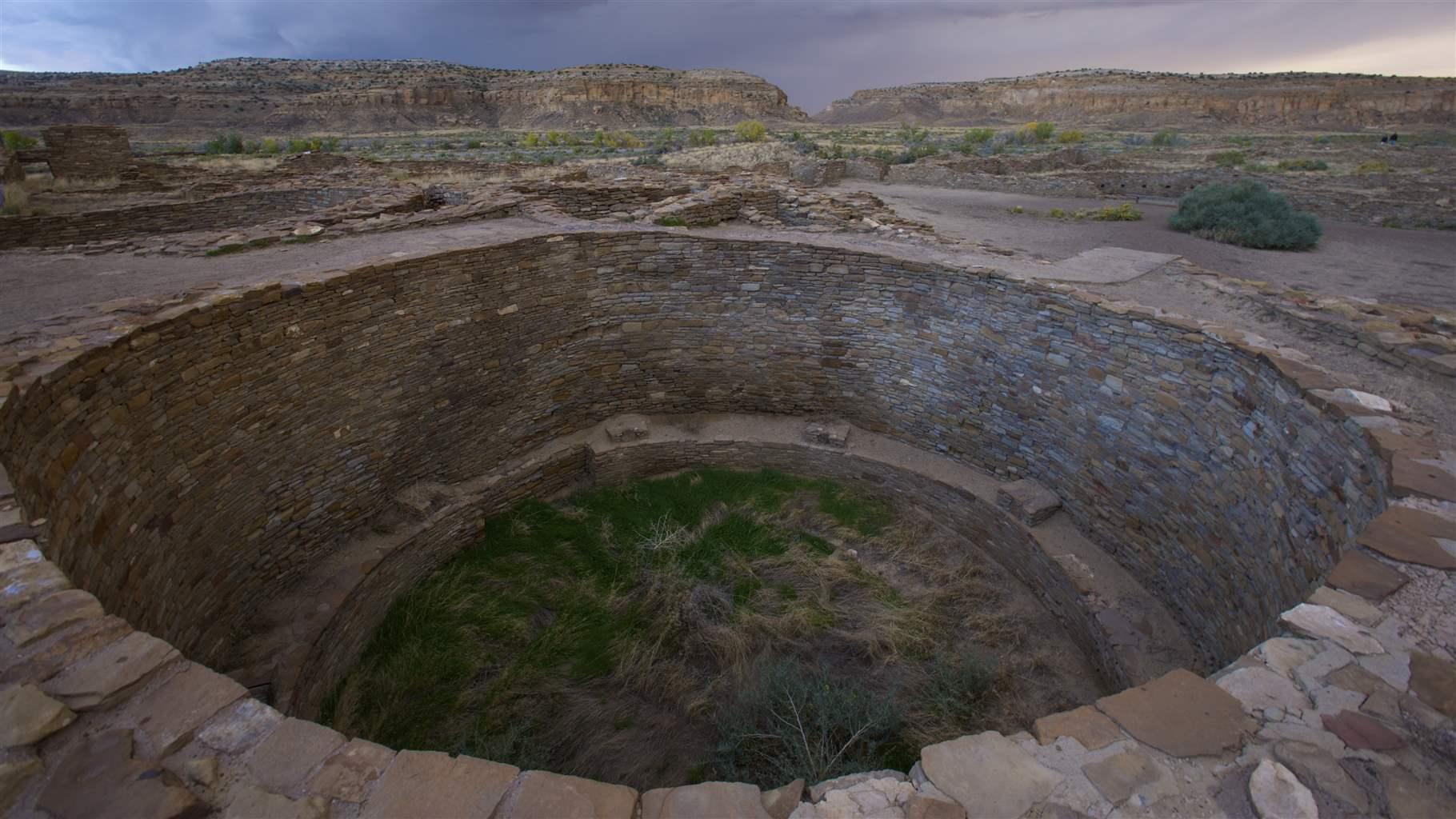 Pueblo Bonito is the largest “great house” that Indigenous people built hundreds of years ago within Chaco Canyon, New Mexico. Landscape view of Pueblo Bonito surrounded by grass a dusk