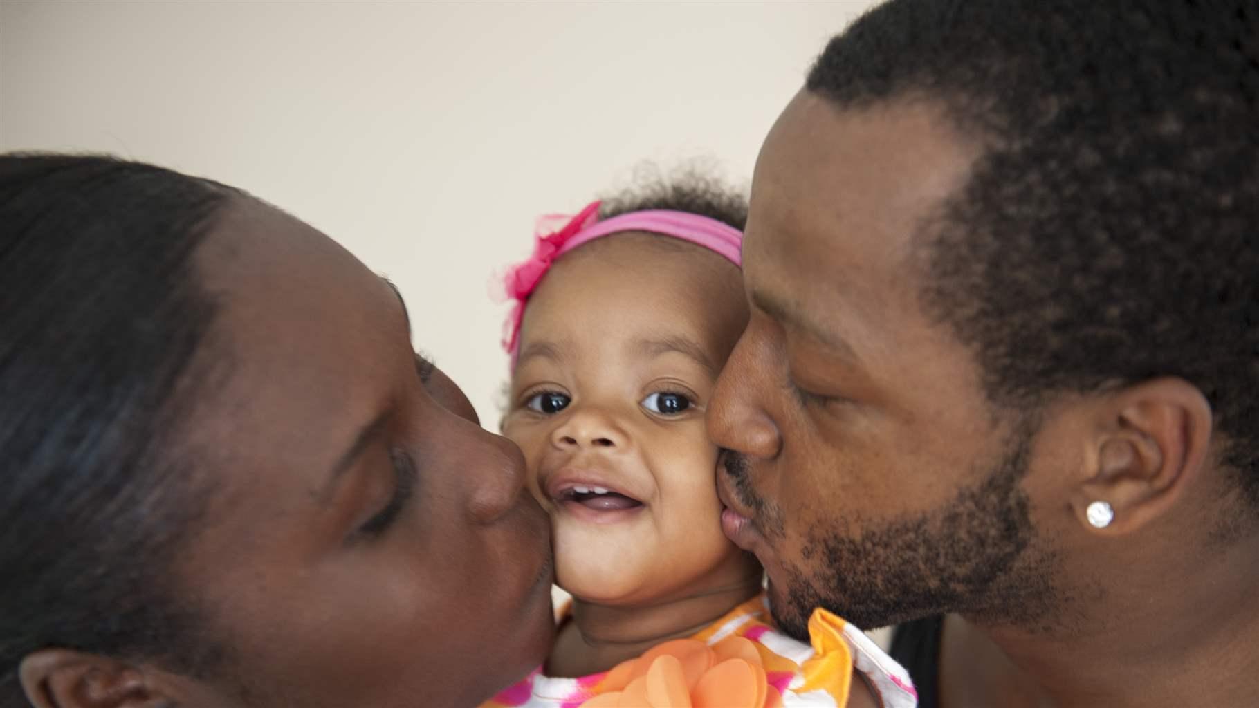 Side profiles of a mother and father kissing the cheeks a baby girl who is in the middle