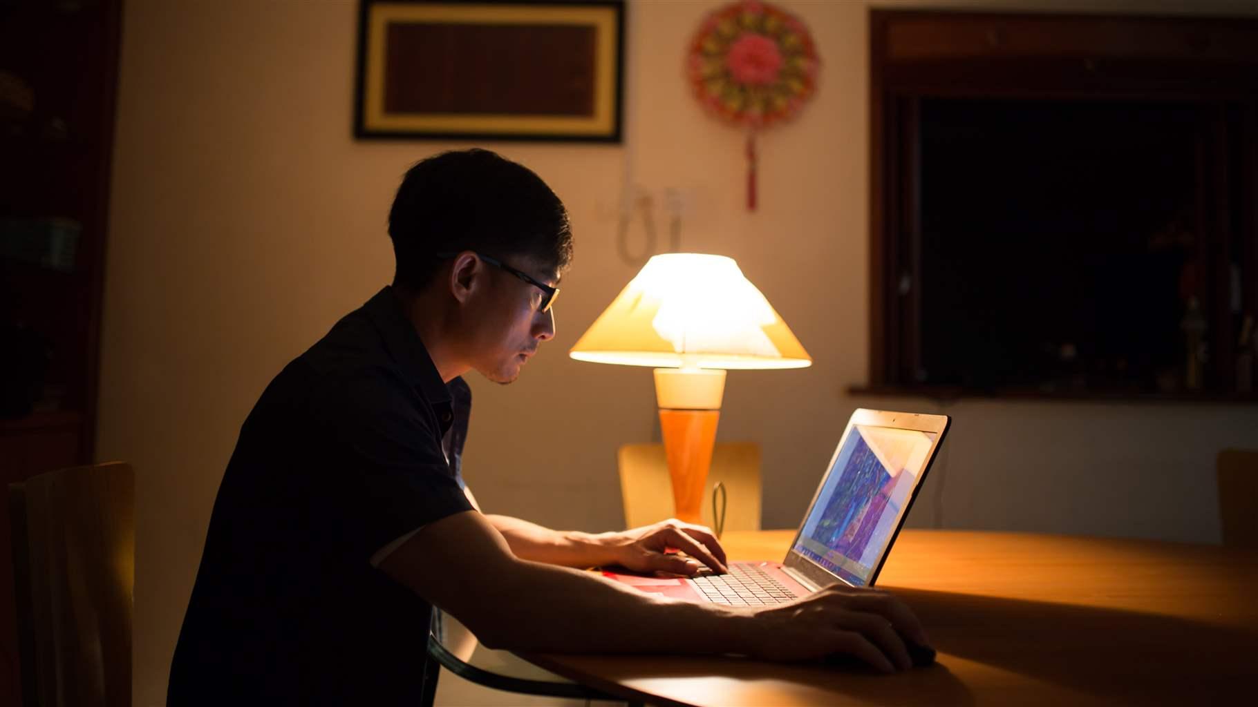 A man sitting at a dinning room table with his laptop and lamp.  
