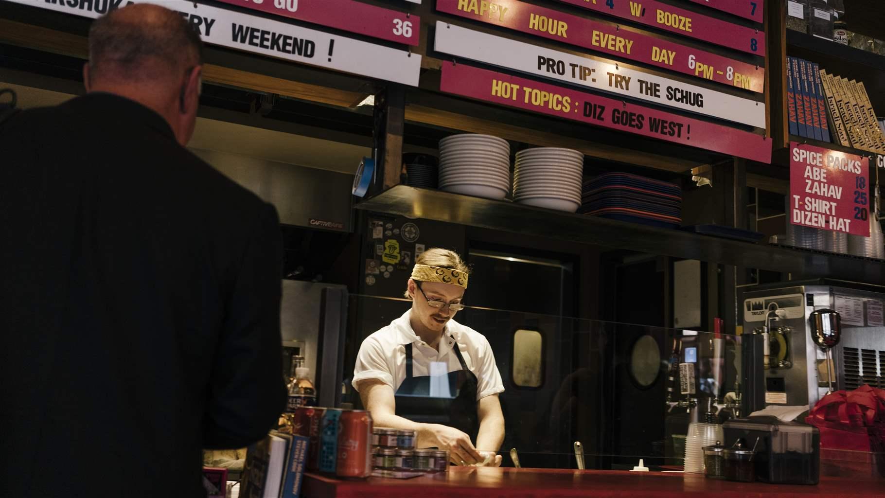 A man behind a counter prepares an order with a customer waiting.