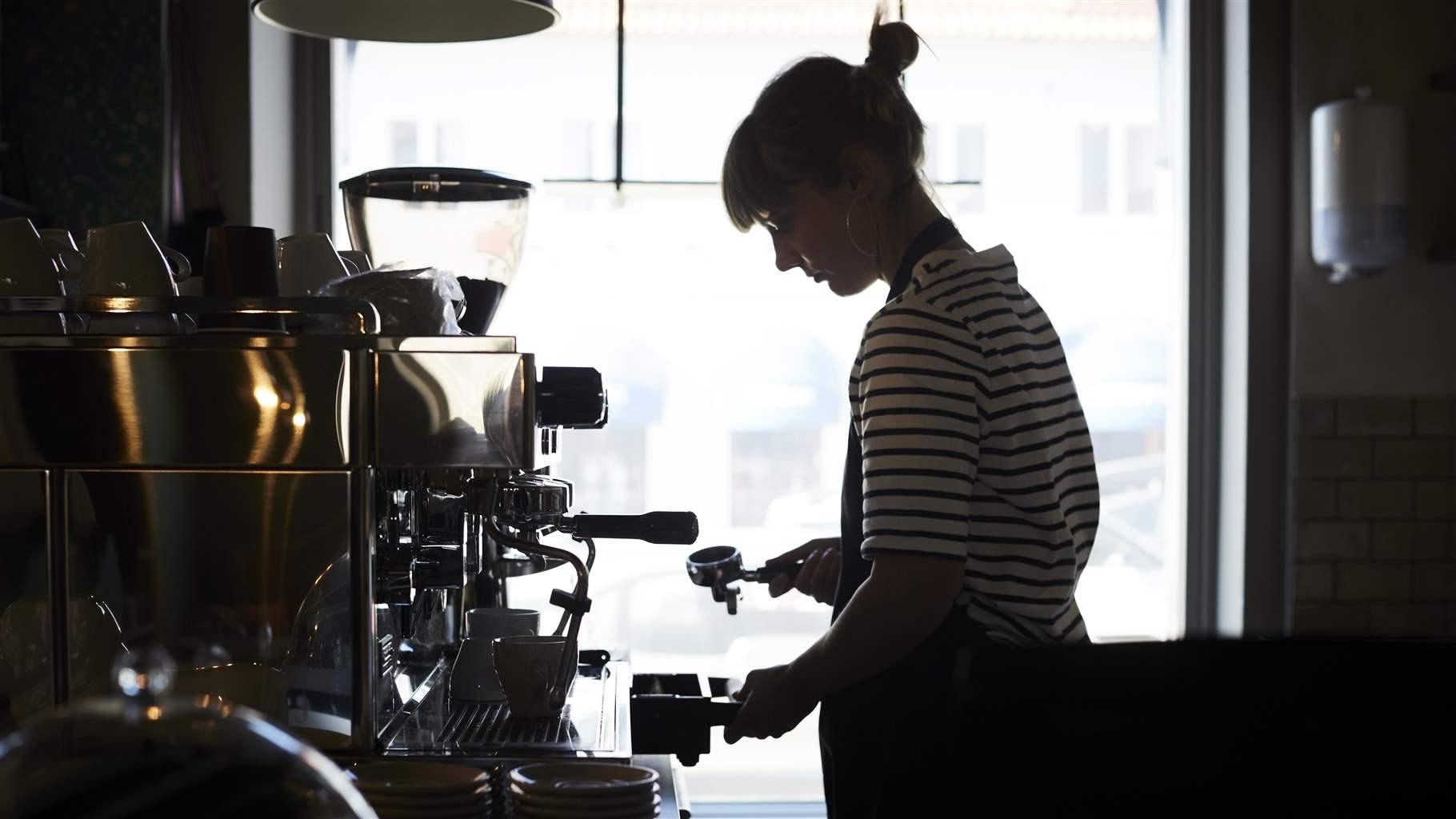 A barista in silhouette as she is working at an espresso machine. 