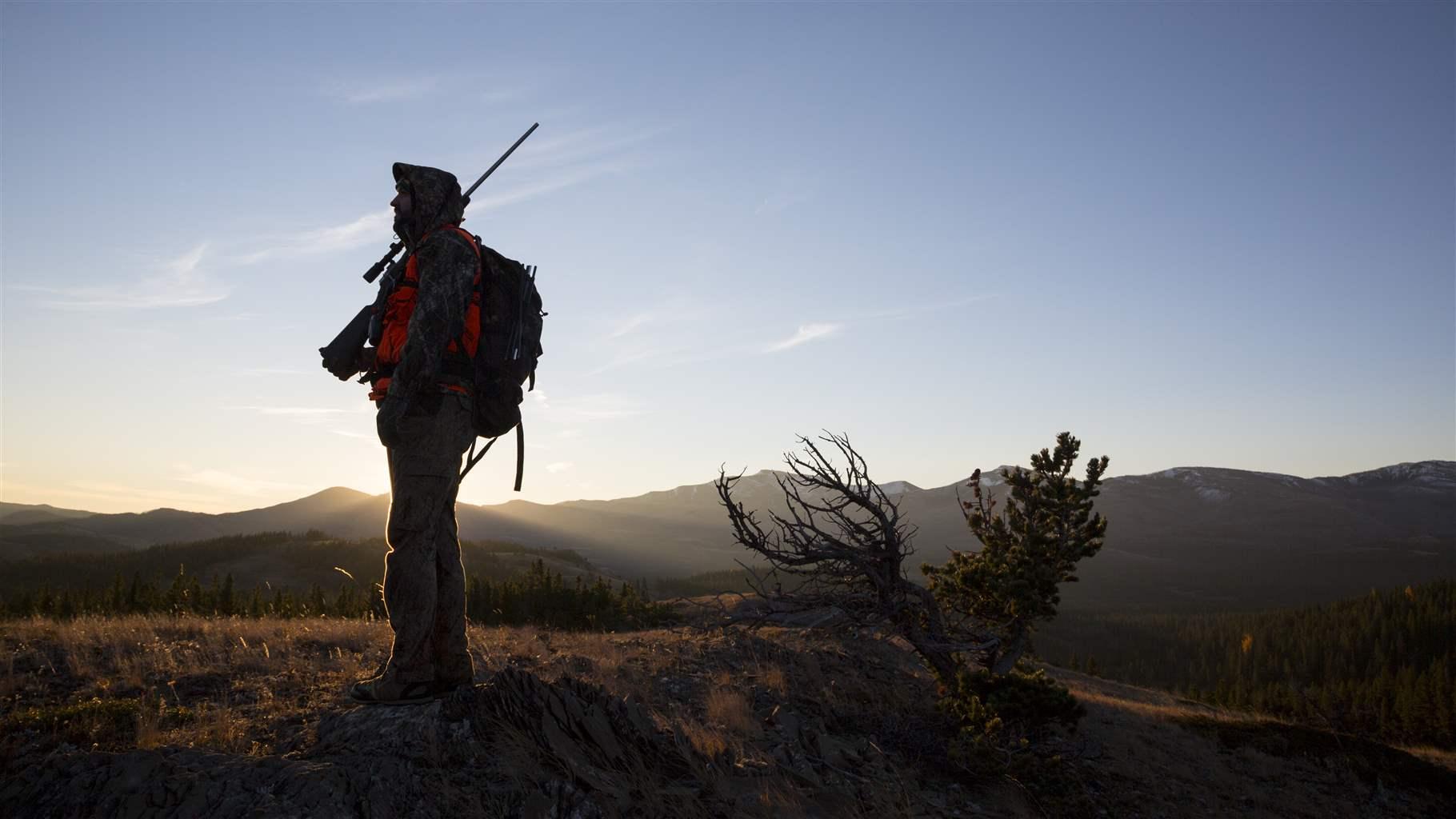 A hunter, carrying a backpack and with a rifle slung over his shoulder, stands in silhouette at dusk with mountains in the distance.