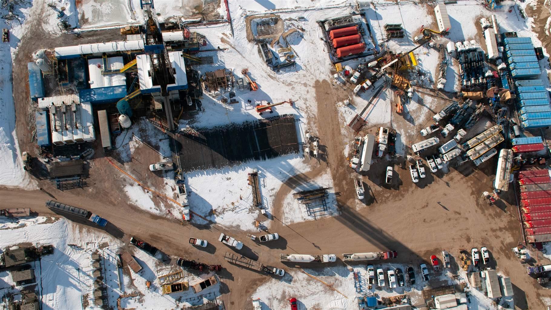 A close aerial view of a well pad, dusted with snow, with vehicles, drills, container boxes, and other equipment densely packed together.