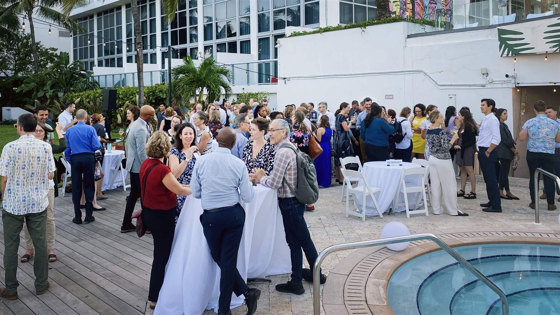 Conference attendees socialize on a patio near a pool. Palm trees and a white building with windows are in the background. 
