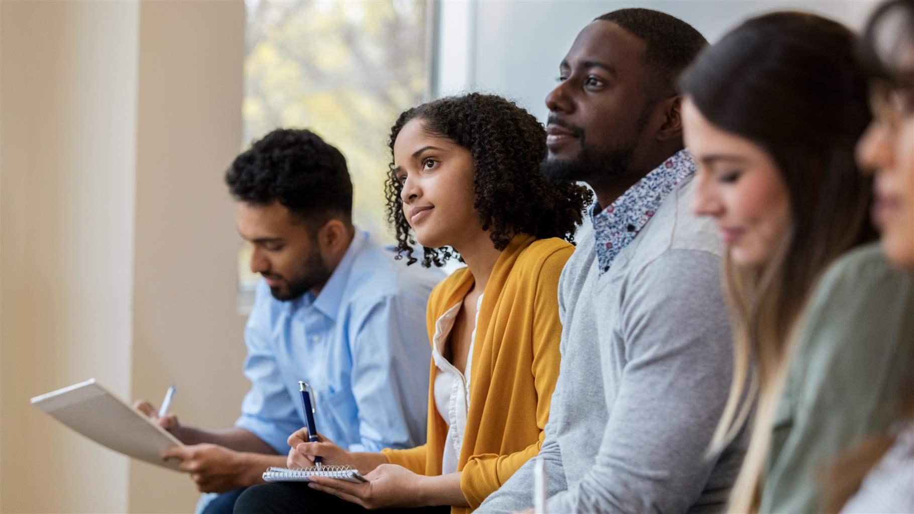 A group of business people sit in a row in a training class. They look at an unseen speaker as they concentrate on his lecture