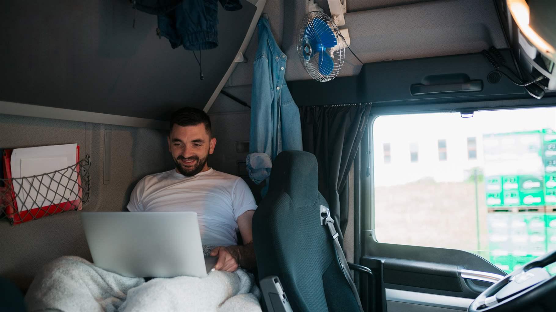 Male trucker sitting in the truck's sleeping cabin, relaxing and using a laptop to make a video call and talk to his family while on the road
