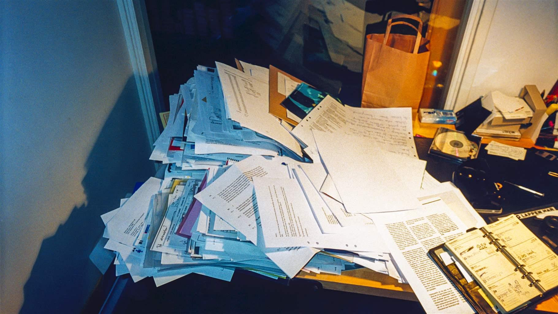 A close-up view of a cluttered desk strewn with large stacks of paper, a day planner, CD-ROM, and other items. The mess casts a gray shadow on the wall to the left and reflects in the window behind the desk.