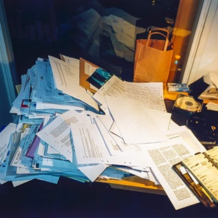 A close-up view of a cluttered desk strewn with large stacks of paper, a day planner, CD-ROM, and other items. The mess casts a gray shadow on the wall to the left and reflects in the window behind the desk.