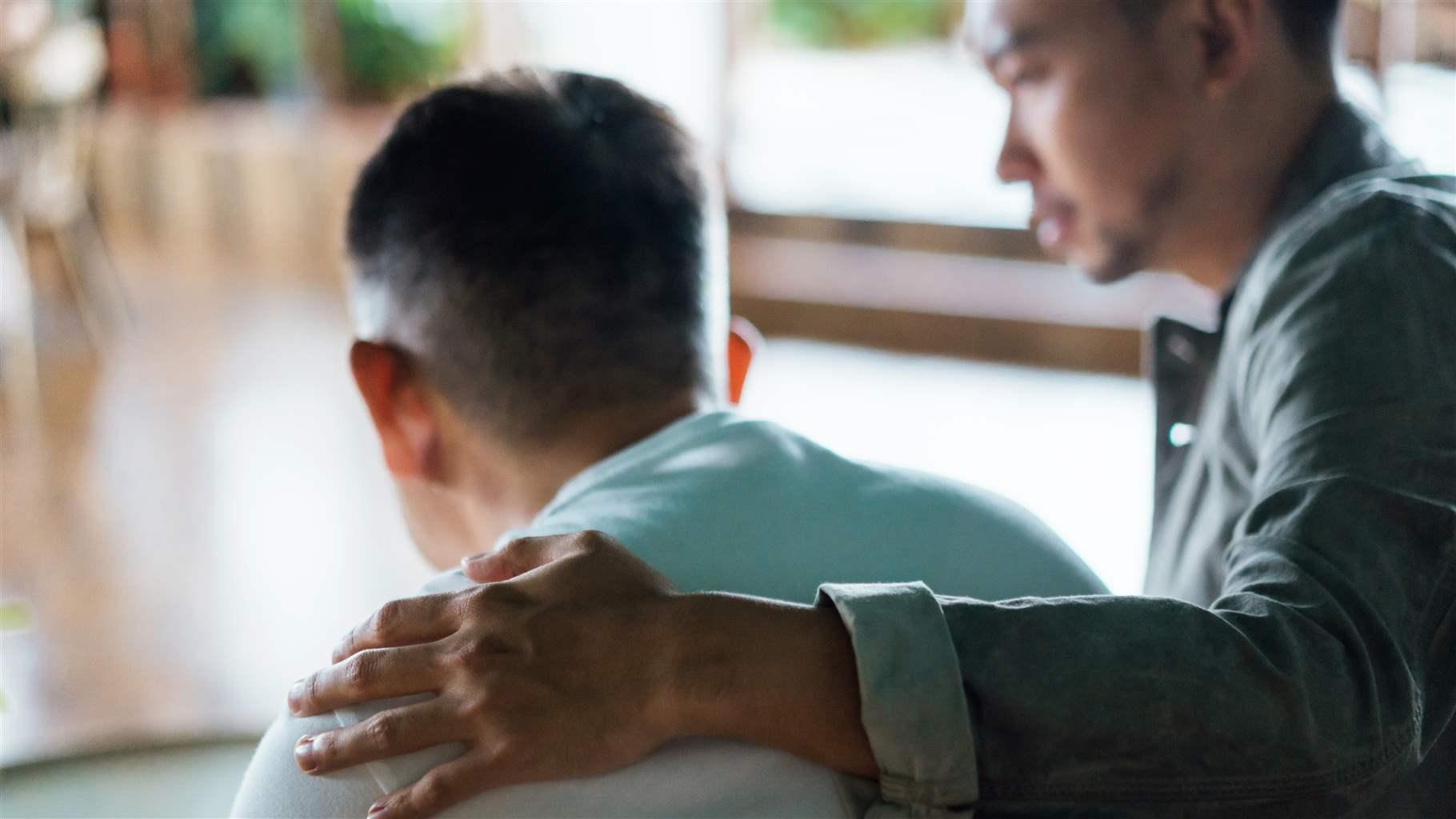 Rear view of son and elderly father sitting together at home. Son caring for his father, putting hand on his shoulder, comforting and consoling him.