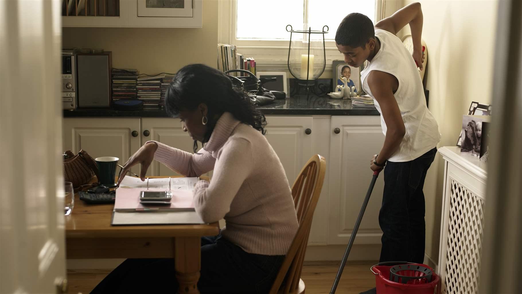 Boy (12-13) mopping kitchen floor behind mother counting bills