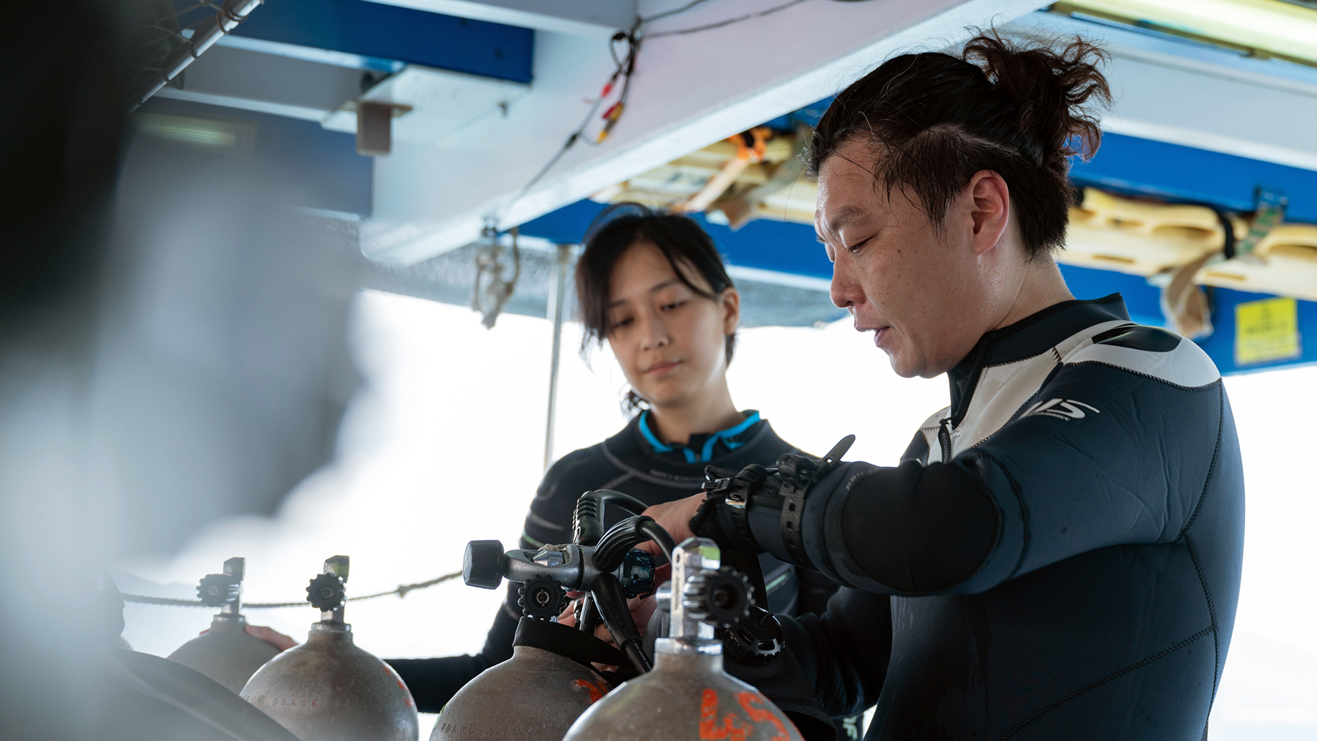 Between a blurred background and foreground, two people in wetsuits come into sharp focus, standing next to a line of tall silver scuba tanks that each have small, bright orange numbers painted on one side.