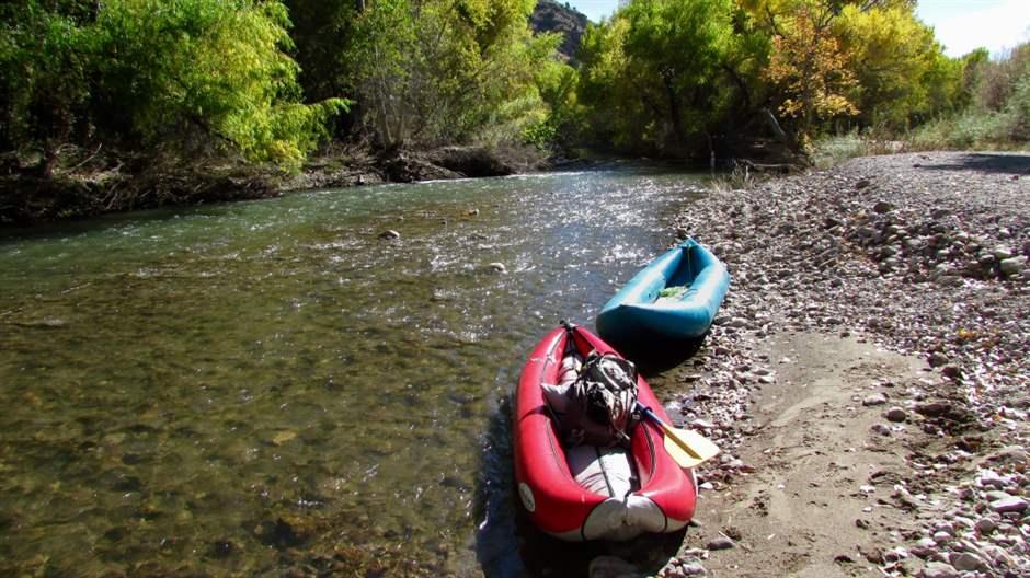  Two unmanned inflatable kayaks—one red and one blue—sit on the shore of a shallow stream lined on one side by dense brush and on the other by a bank of small, loose stones and a patch of sand. The sky is sunny and clear.