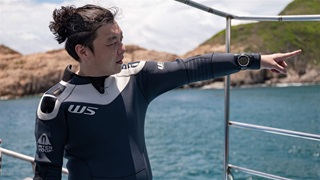 佘国豪穿着潜水衣站在一条船上，身后是波光粼粼的蓝色海水和丘陵海岸，他在潜水前指向远处的水域。