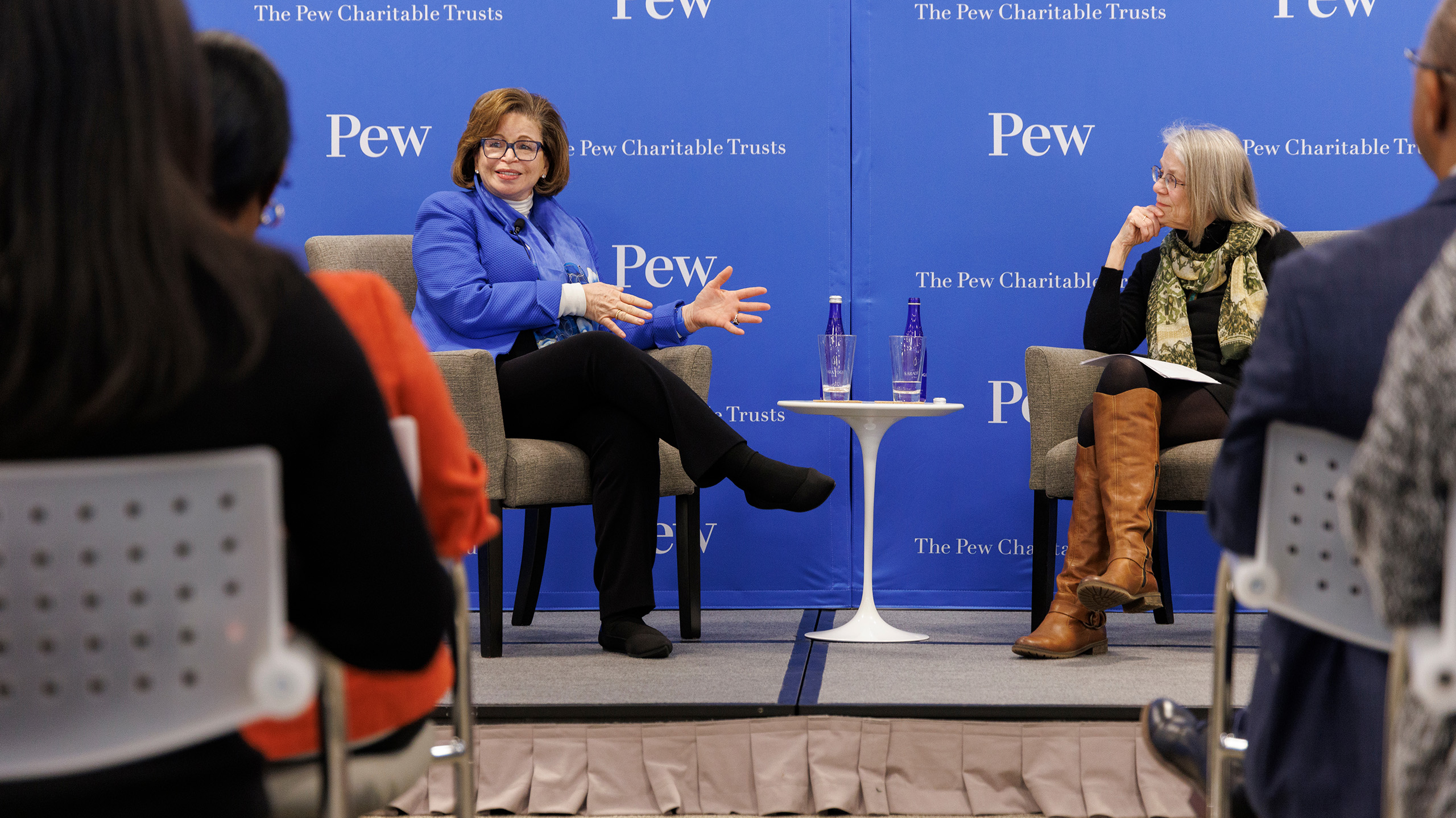 Valerie Jarrett and Susan Urahn sit facing each other on stage, in front of a room filled with Pew staff members. Jarrett and Urahn are seated in tan upholstered chairs on opposite sides of a small white pedestal table, in front of a blue backdrop featuring the Pew logo.  