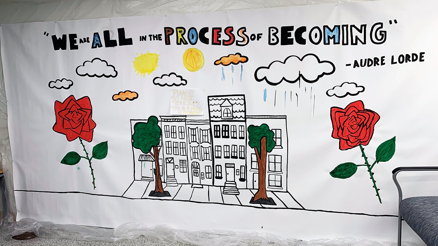 The mural is painted on a large roll of white paper and features a quote from writer Audre Lorde: “We are all in the process of becoming.” It features a black-and-white sketch of a block of row houses framed by two red roses, with clouds and the sun overhead. 