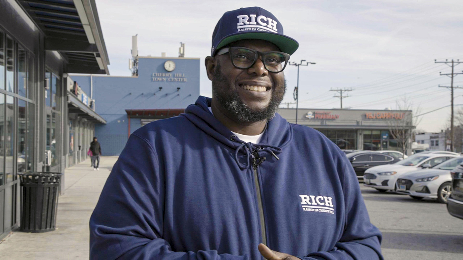 RICH co-founder Michael Battle stands in front of the Cherry Hill Town Center. He has a full beard, glasses, and a big smile, and is wearing a cap and a navy blue sweatshirt, both emblazoned with the RICH logo.
