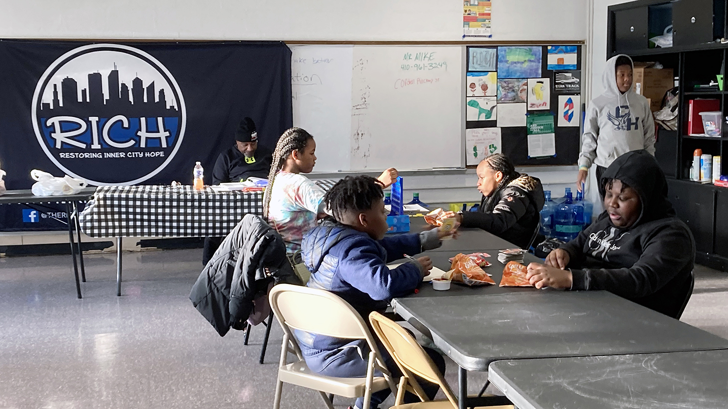 Four students sit at long tables playing games and eating snacks in RICH’s after-school program room, decorated with a large RICH banner that features an outline of the Baltimore skyline.